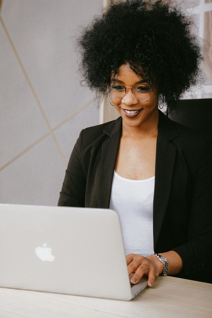Smiling woman with afro hair working on a laptop in a modern office setting.